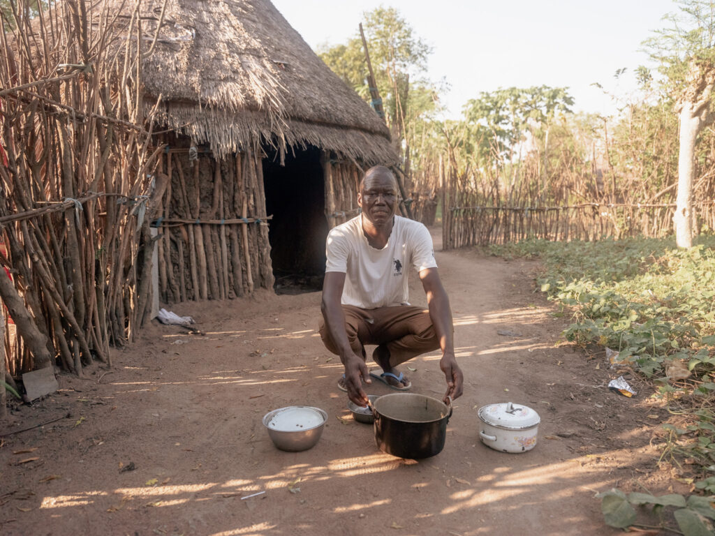 Man kneling in front of hut with a couple of pots.