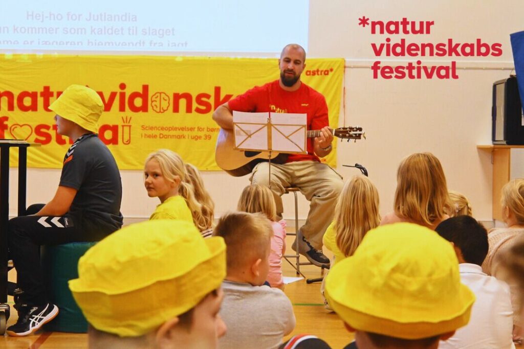 Mand der spiller guitar iført rød t-shirt omgivet af børn i gule naturvidenskabsfestival-bøllehatte.