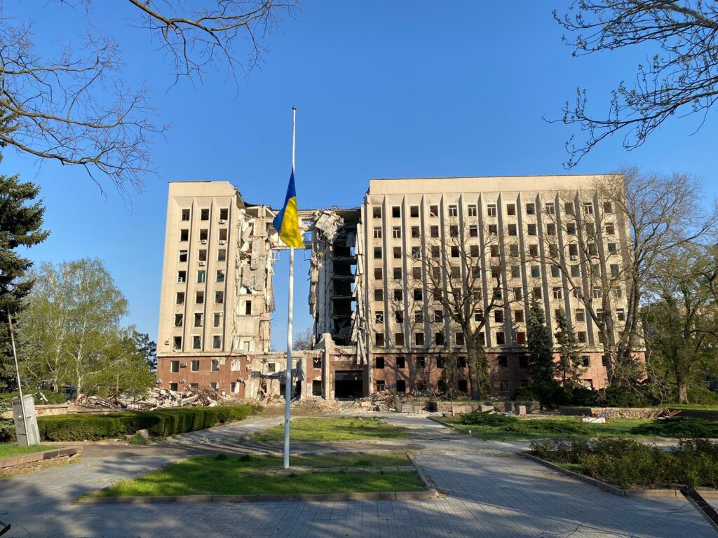 Building damaged by shelling behind a flag pole with the Ukrainian flag.